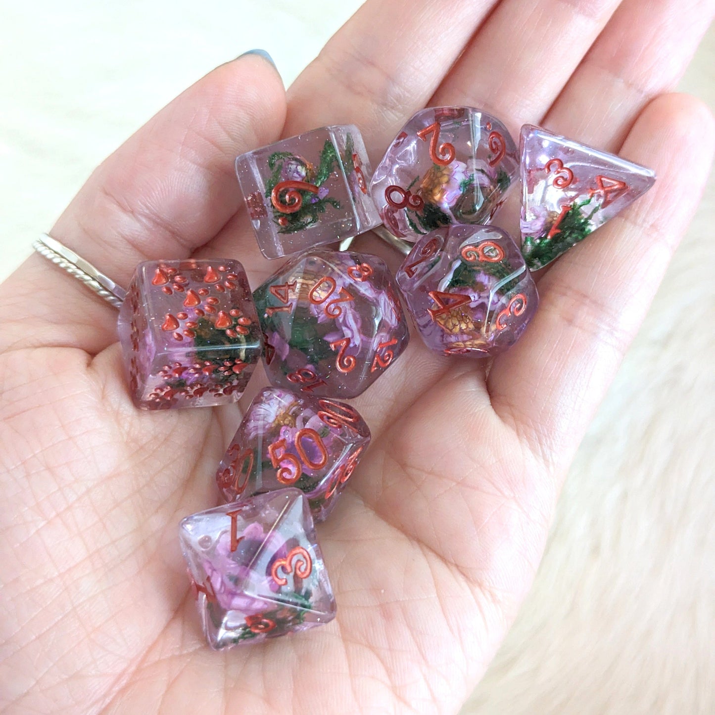 Hand holding purple flowers and moss dice set with coppery red font, featuring real dried flowers in tinted purple resin with glitter.