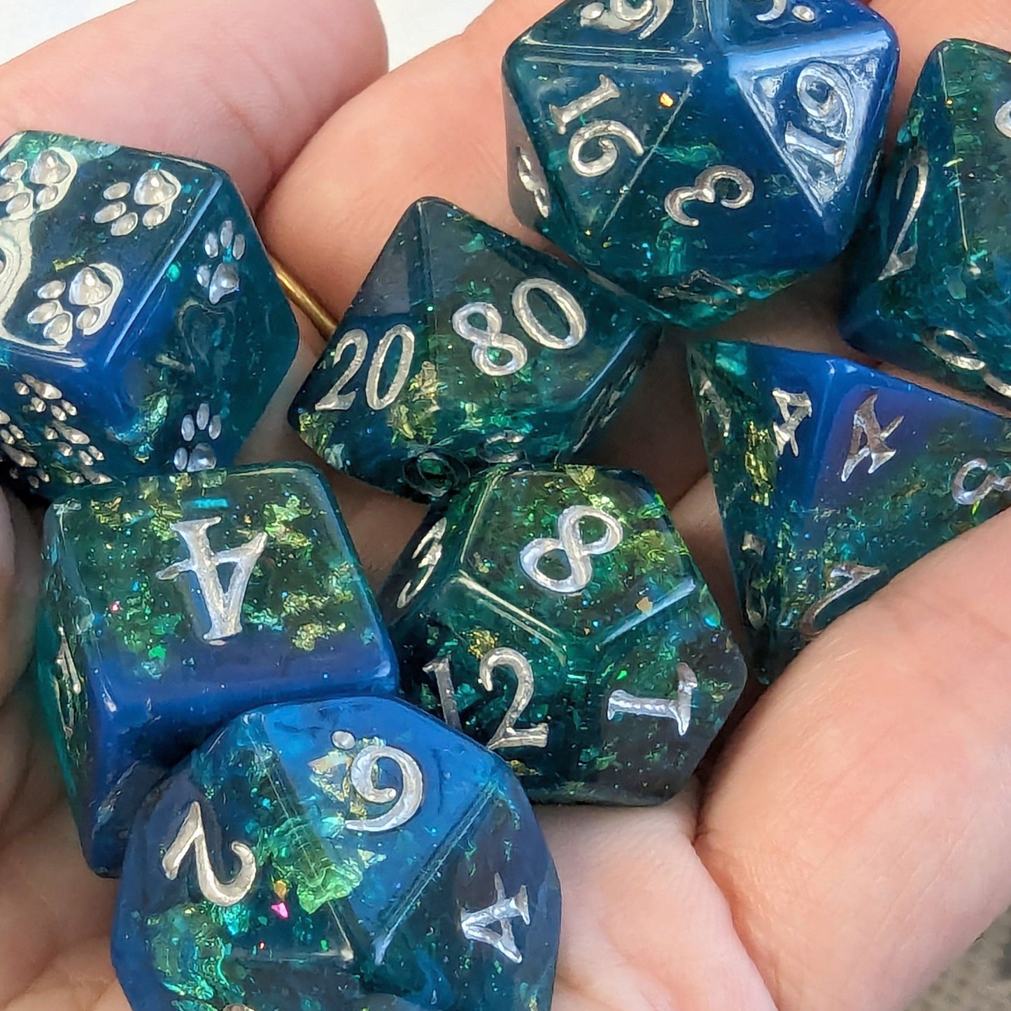 Hand holding Saltmarsh Dice Set with blue and green opal flakes, gold foil, and paw design D6, perfect for Dungeons and Dragons.