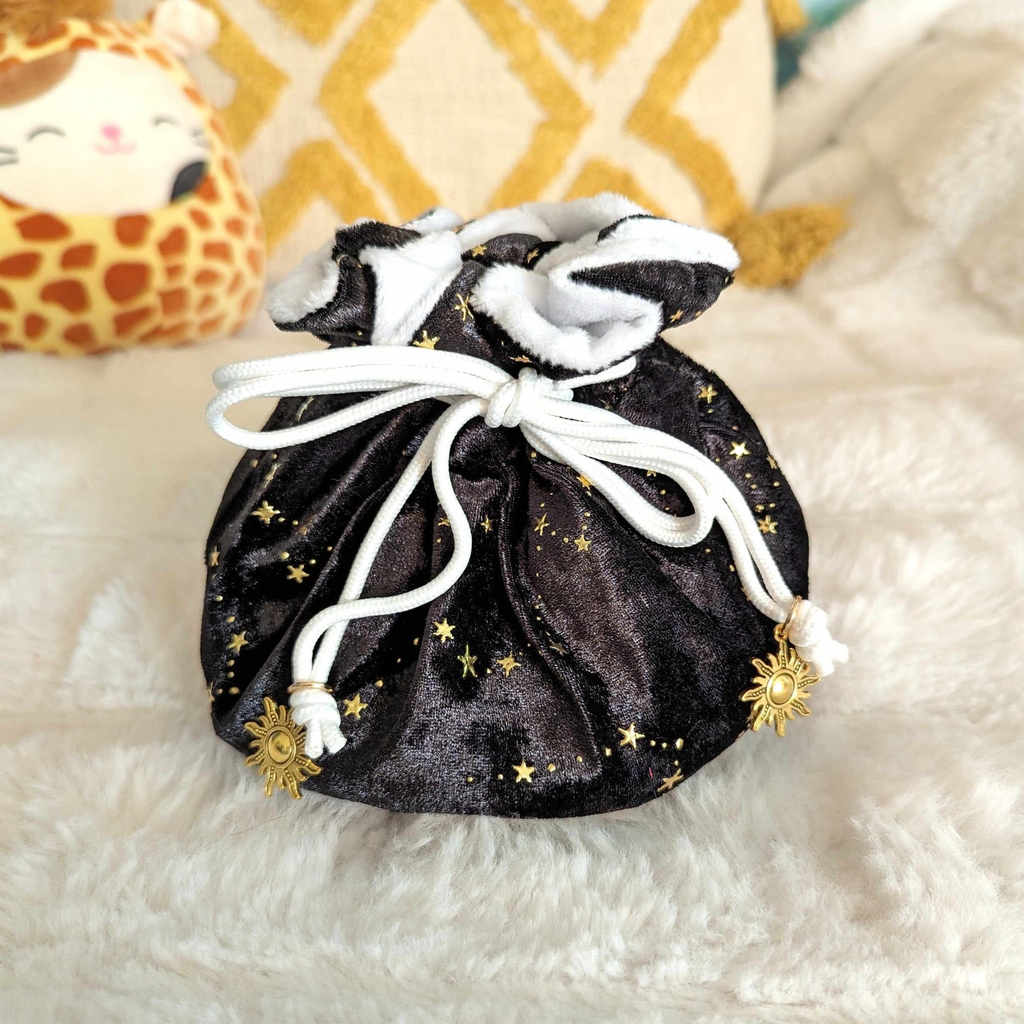 Black velvet dice bag with gold stars, white drawstrings, and sun charms on a plush white surface.