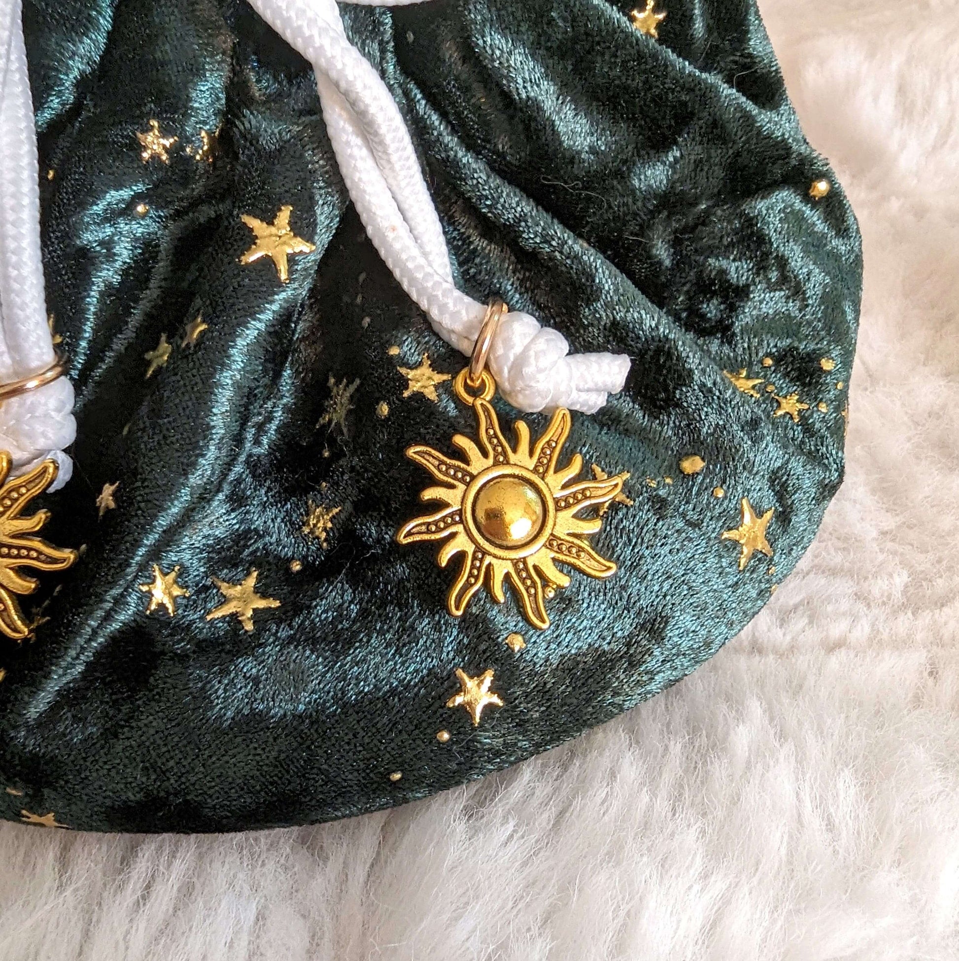 Luxurious green velvet dice bag with golden star patterns and elegant sun charm accents on white drawstrings, resting on soft white fabric.