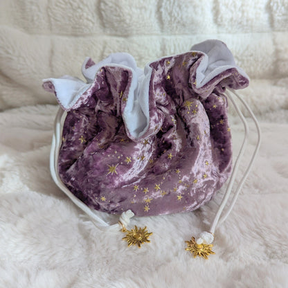 Starry mauve velvety dice bag with gold stars and solar charms, featuring white drawstrings on a soft fabric background.