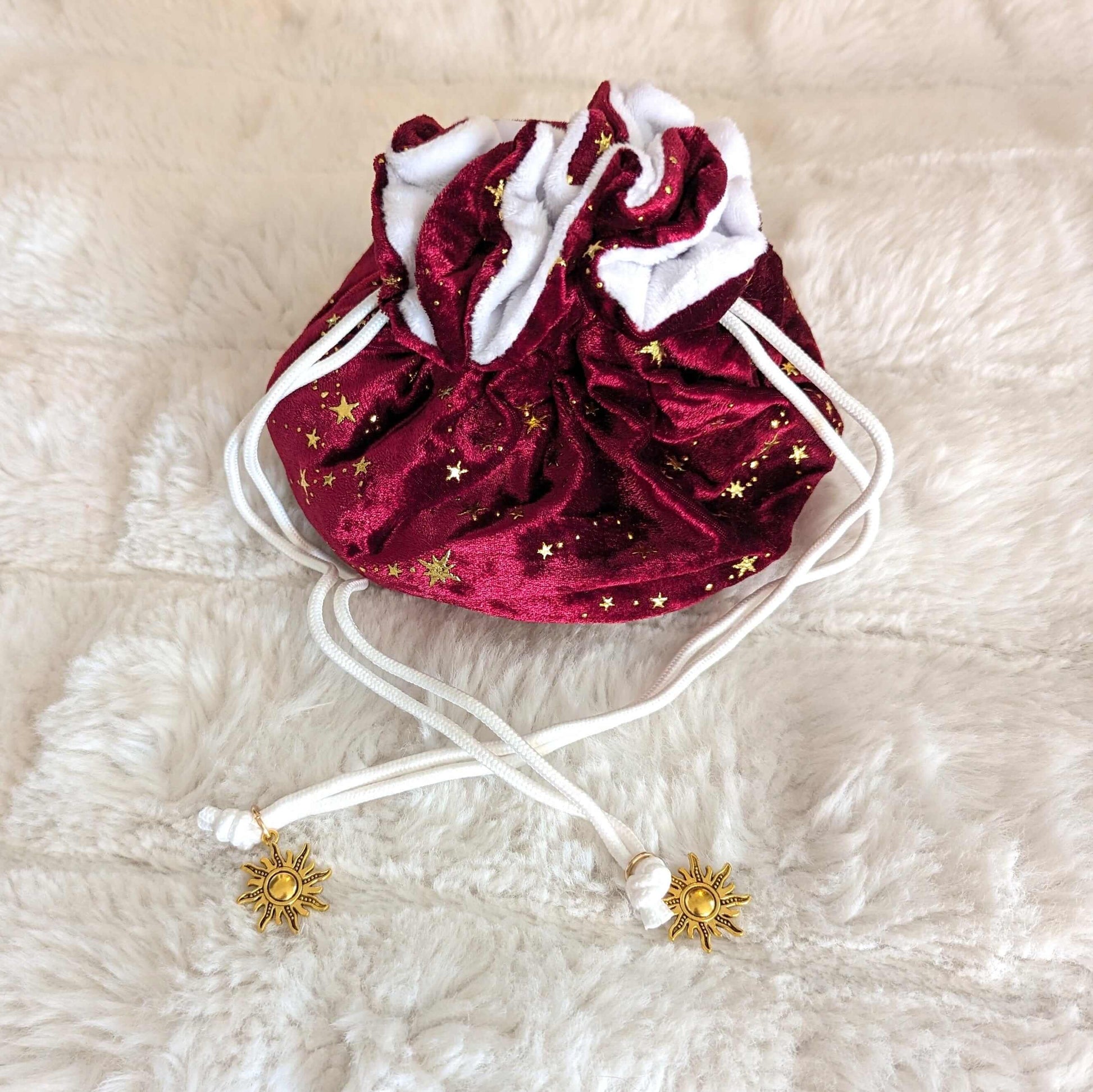 Luxurious red velvet dice bag with golden stars, white plush lining, and elegant drawstrings with golden sun charms, on soft fur background.