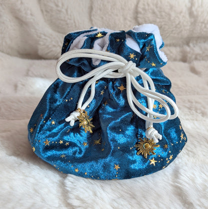 Starry teal dice bag with gold stars, white cord drawstrings, and gold solar charms on a soft fabric background.