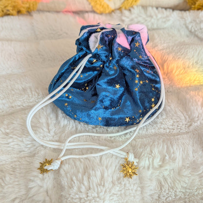 Starry teal dice bag with gold stars and solar charms on a drawstring, resting on a plush white surface.