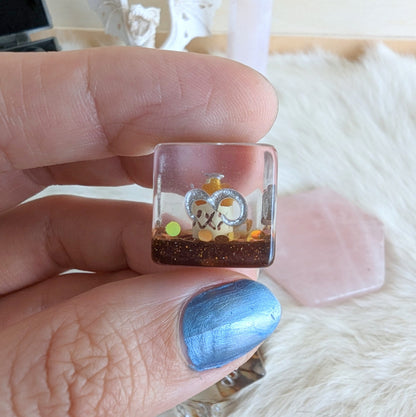 A hand holding a transparent die with decorative elements inside, including a small jug and glitter, part of a Tavern Quest Dice Set.