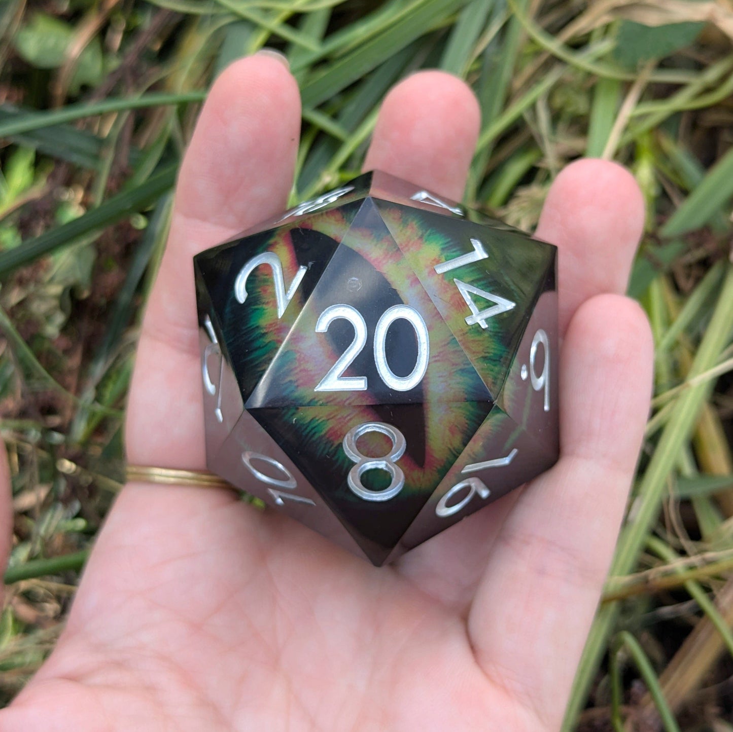 "The Infernal Watcher Giant D20 die with vivid eye design and dark resin exterior, held in hand outdoors."