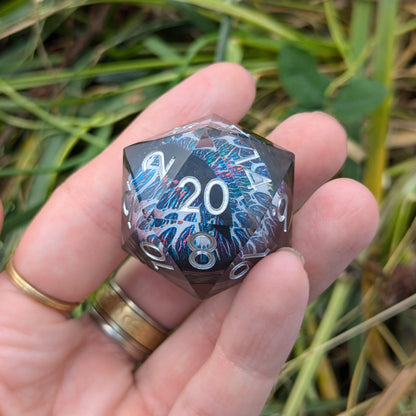 Hand holding the Watchful Eye D20 Clear Resin Edition with white, blue, and black eye detail against a nature background.