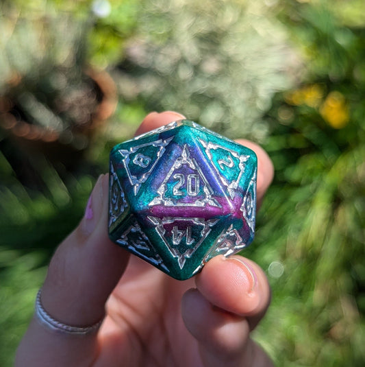 Hand holding a large 20-sided purple and green glitter dice from the Underwater Castle Dice set against a blurred natural background.