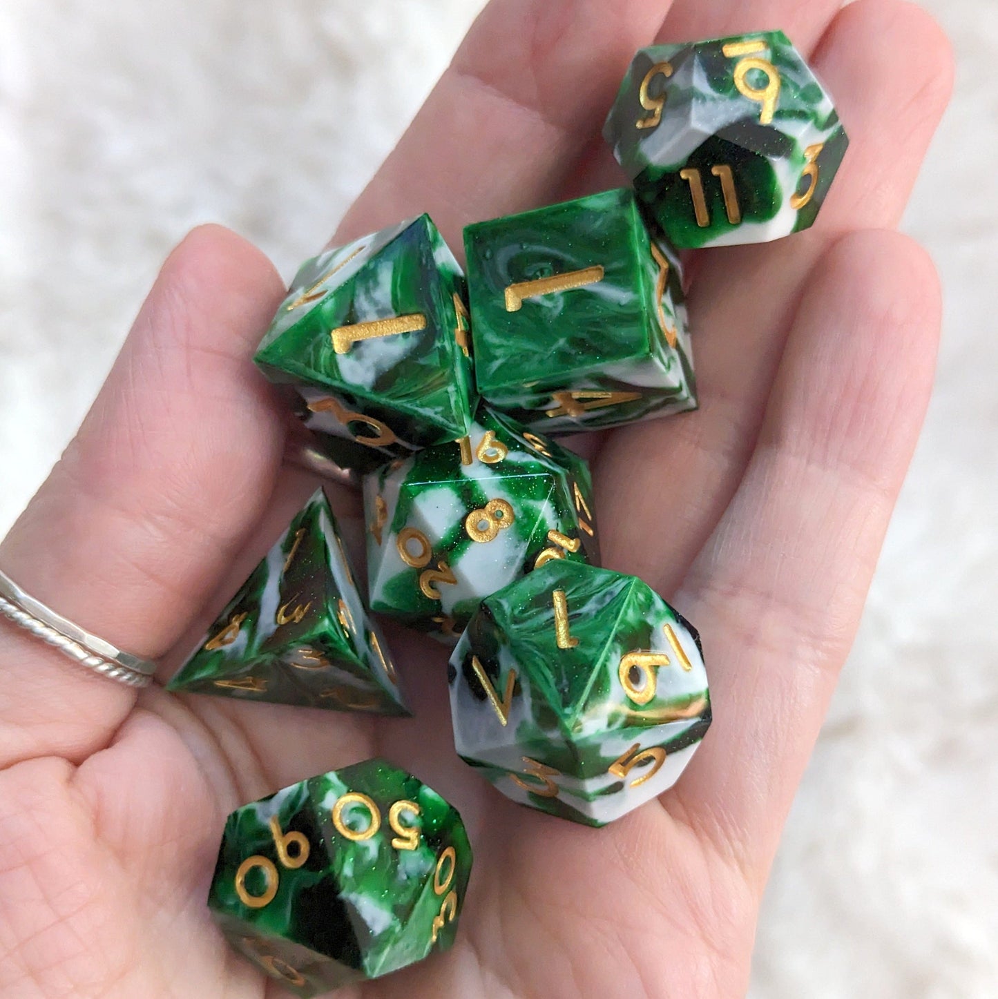 Green and white marbled 7-piece resin dice set for Dungeons and Dragons, featuring gold numbers, held in a hand on a soft background.