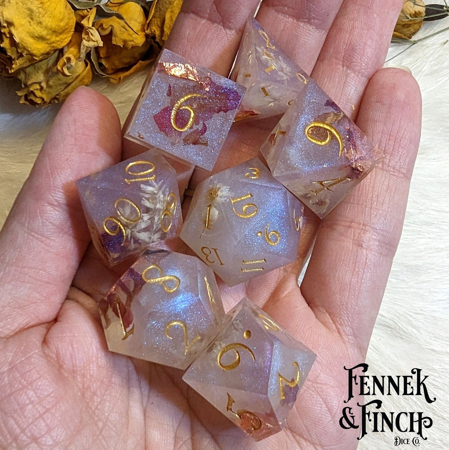 Hand holding sharp edge resin dice set with daisies and rose petals, gold numbers, designed by Fennek and Finch for D&D and Magic games.