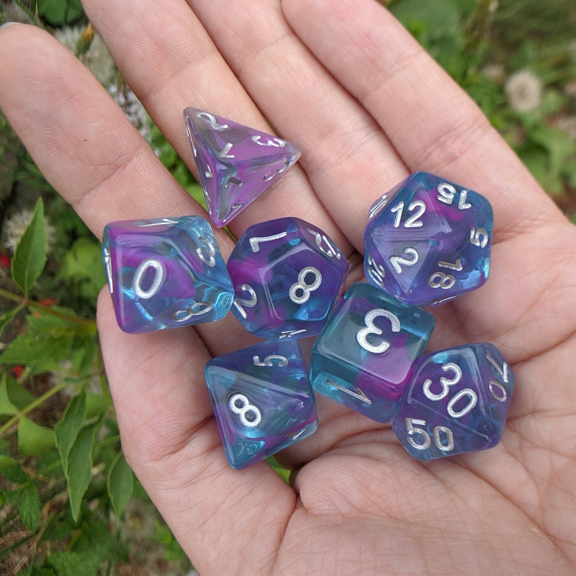 Hand holding Aquarium Ribbon dice set, clear blue and purple inked DnD dice, outdoors with greenery background.