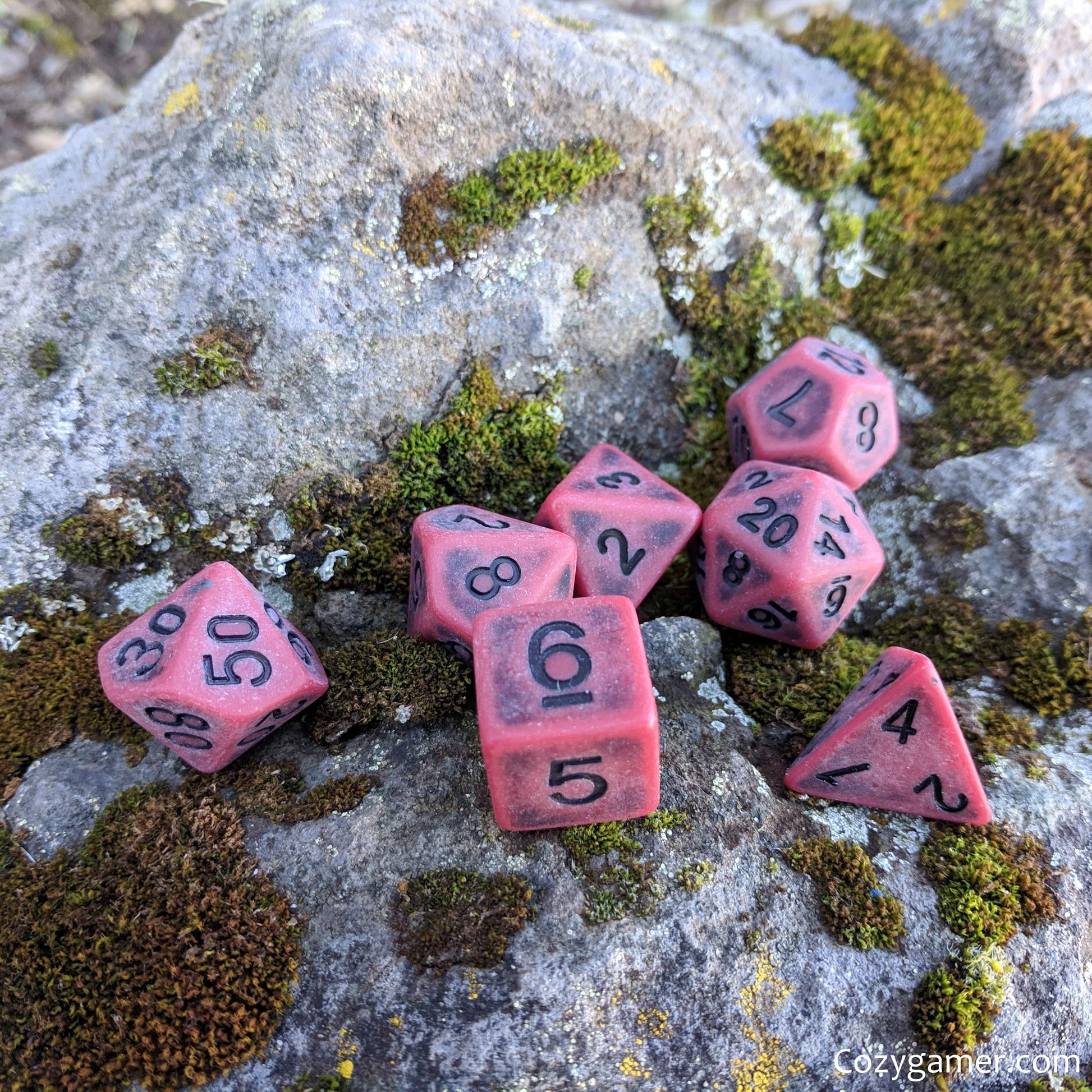 Matte red Blood DnD dice set on mossy rock, 7 polyhedral pieces for tabletop RPGs like Dungeons and Dragons.