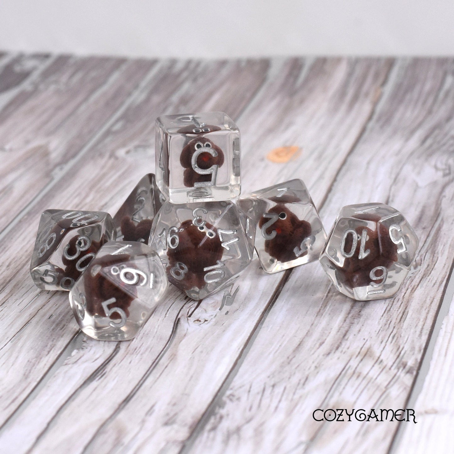 Clear resin dice set with tiny brown dogs inside for tabletop RPGs, 7-piece polyhedral set by CozyGamer on wooden surface.