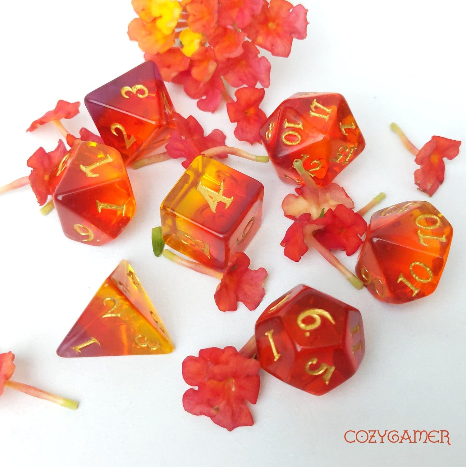 Dragon Fire Dice Set in ombre shades of purple, red, orange, and yellow with pale gold numbers, surrounded by red flowers.