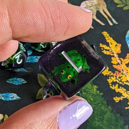 Close-up of Hoppy Hauntings Frog DnD Dice Set featuring a green frog inside a clear purple die, perfect for magical tabletop games.