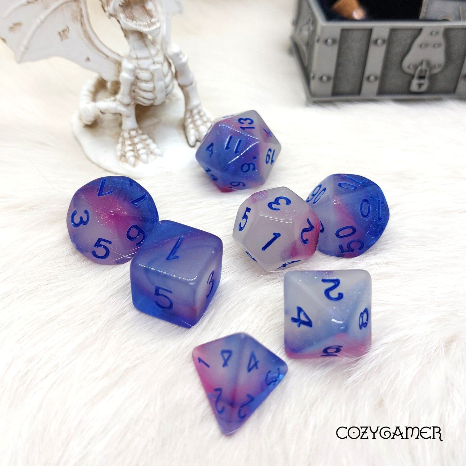 Glow in the dark white, blue, and purple dice set on a white surface with a decorative dragon and treasure chest background.