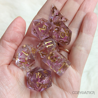 Mauve Immortal Love 7-piece resin dice set held in hand with petals and metal foil for TTRPG games.