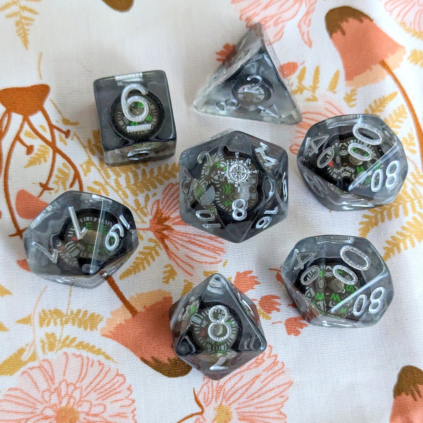 Mini compass 7-piece dice set for DnD, featuring real compasses inside, on floral background. Essential for tabletop games.