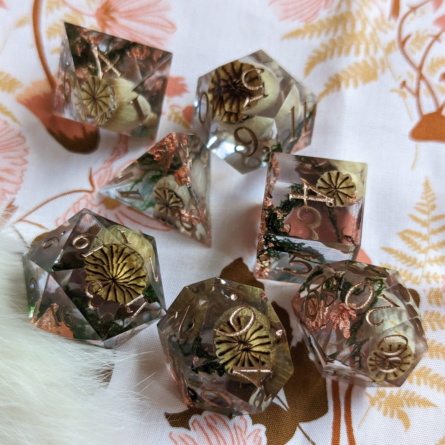 Poppy Pods and Moss dice set featuring poppy pods, moss, and copper foil with gold font, on a floral background.