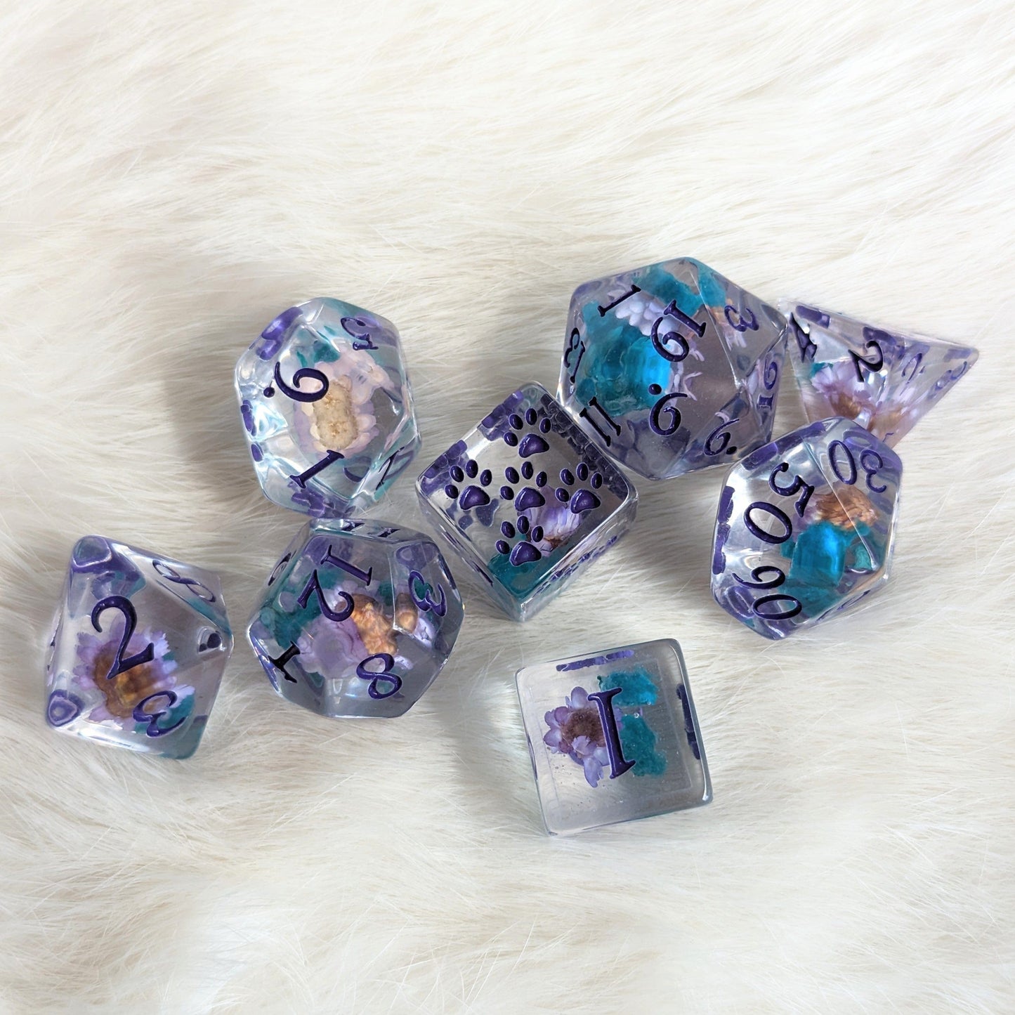 Purple and blue flower DND dice set featuring polyhedral dice with fantasy font and paw-marked D6 on a white fur background.