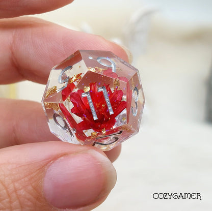 Hand holds polyhedral dice with red flower and gold foil inside clear resin, part of a sharp edge 7-piece DND dice set.