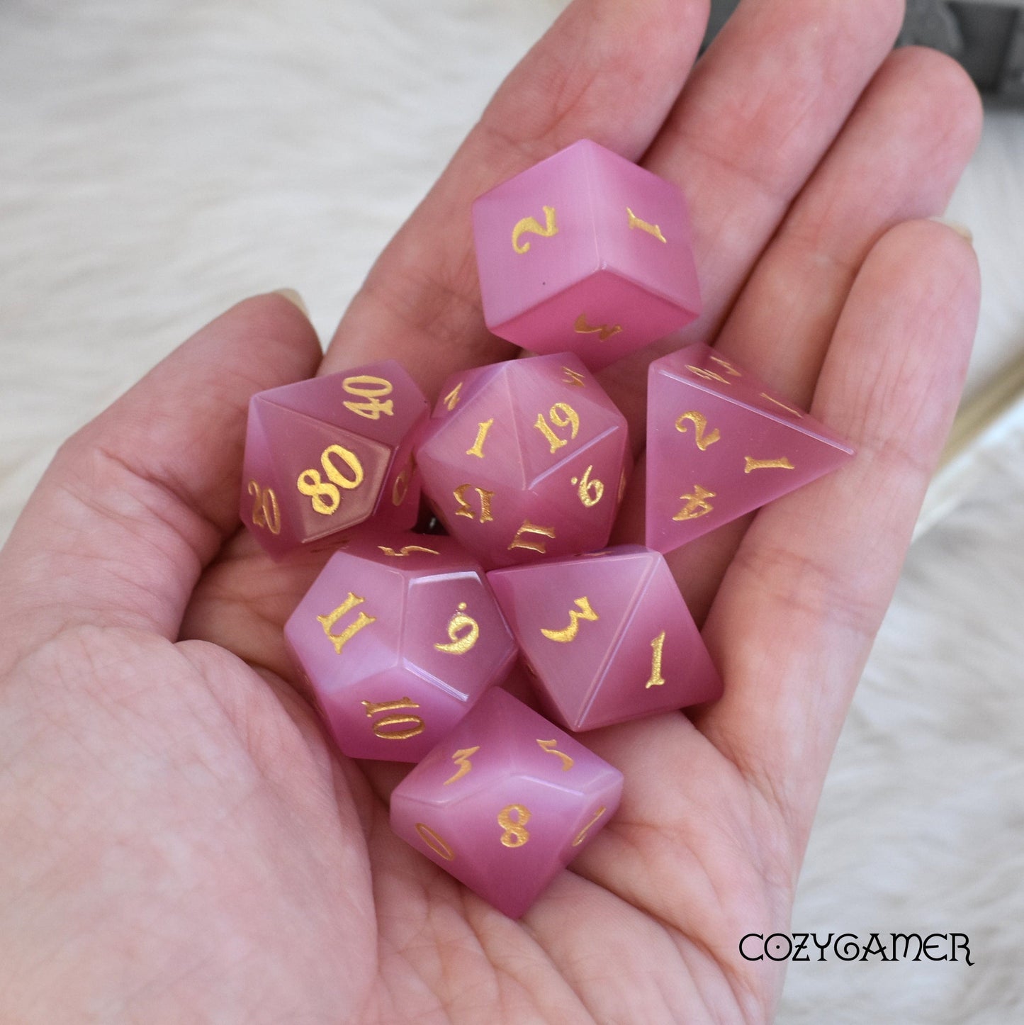 Hand holding a Rose Cat's Eye gemstone 7 piece dice set with gold numbers, perfect for DnD gaming.