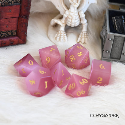 Rose Cat's Eye gemstone dice set for DnD, 7 pink carved stones with gold numbers, perfect gift for gamers, on a fluffy background.