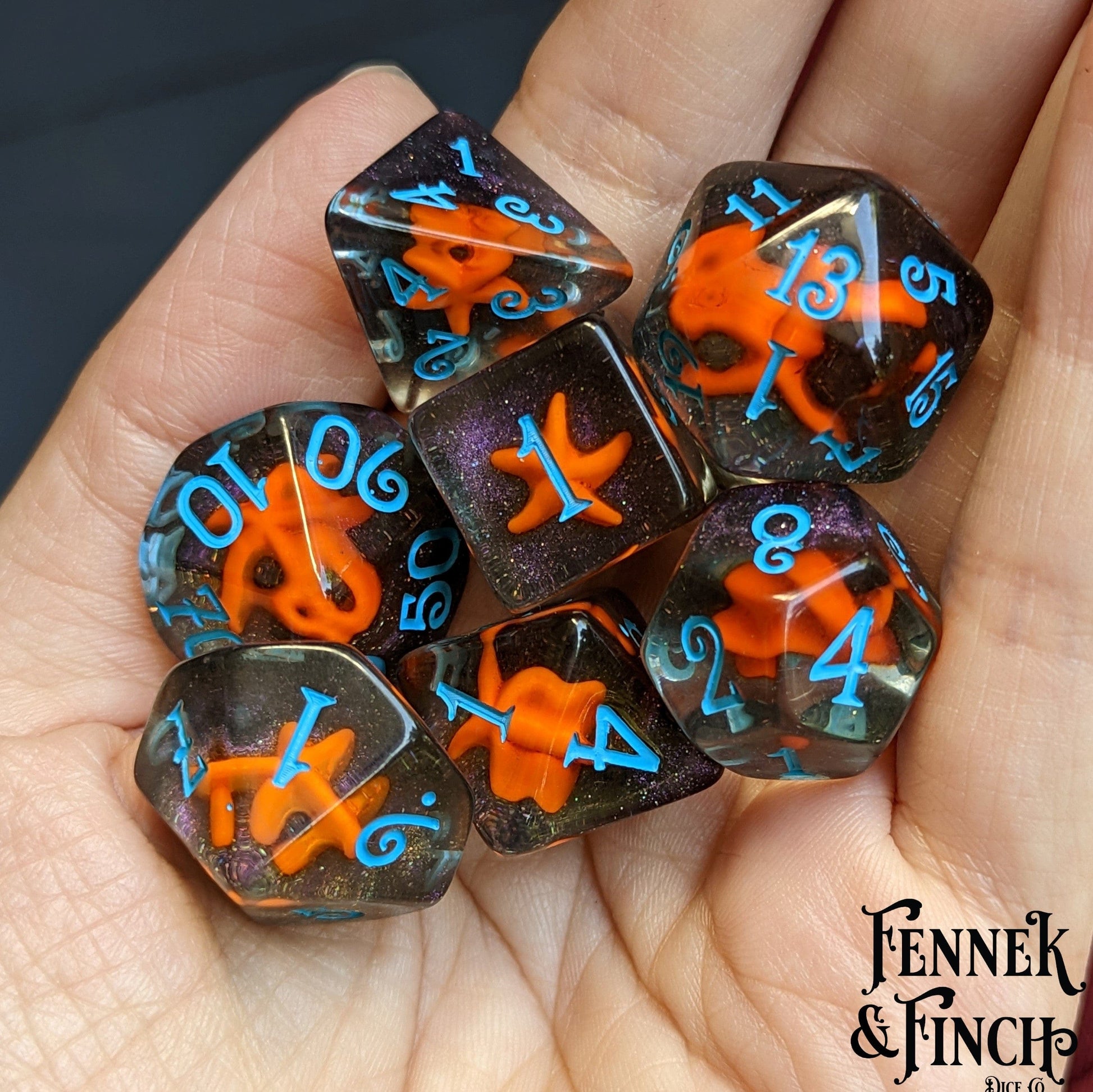 Hand holding 7-piece polyhedral Starfish Dice Set with orange starfish design on black sand, ideal for DND and TTRPGs.