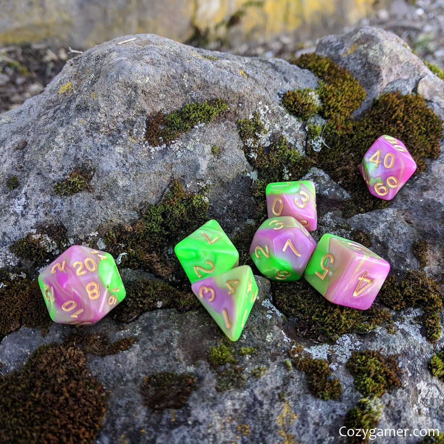 Marbled green and pink polyhedral dice set for tabletop RPGs on a mossy rock background.