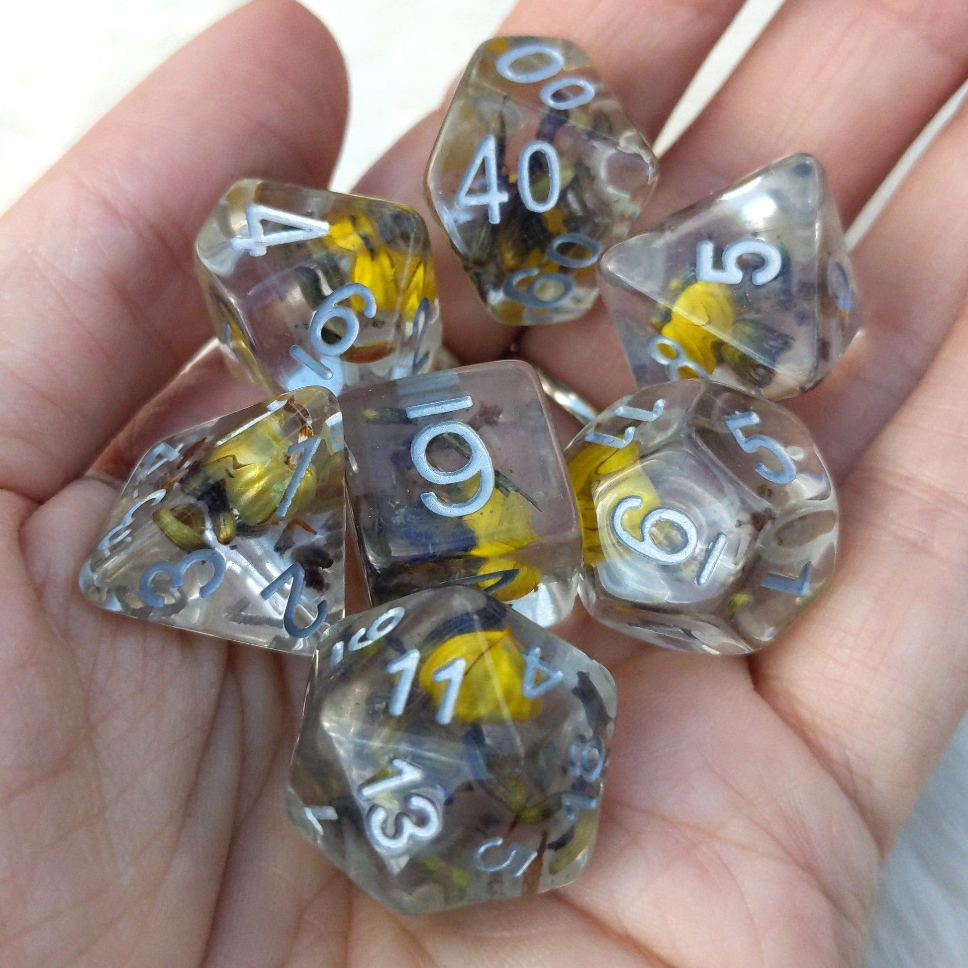 Hand holding a 7-piece polyhedral dice set with real yellow flowers and lavender in clear resin, featuring silver numbers for D&D.